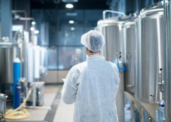 Tecnologo em traje de proteção branco caminhando pela linha de produção da fabrica de alimentos verificando qualidade