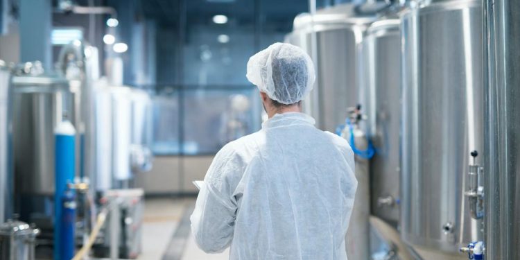 Tecnologo em traje de proteção branco caminhando pela linha de produção da fabrica de alimentos verificando qualidade