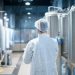 Tecnologo em traje de proteção branco caminhando pela linha de produção da fabrica de alimentos verificando qualidade